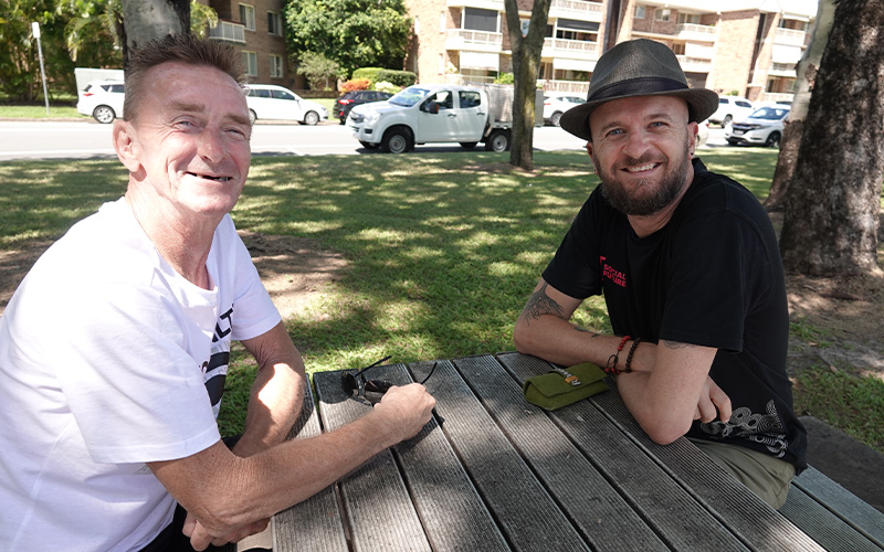 Two men sitting at a picnic table under a tree