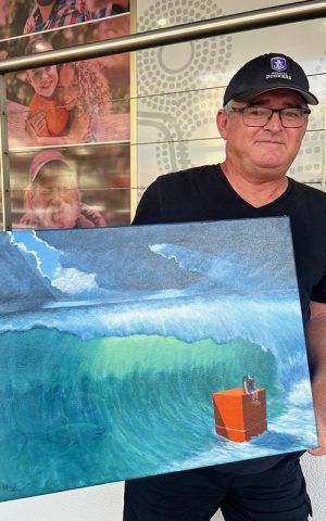 Man wearing a cap and glasses holding a painting depicting a crashing wave