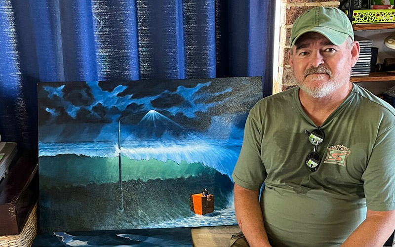 Bearded man in cap sitting in front of a seascape painting