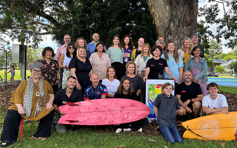 Group of young people, dignitartes and program workers under a fig tree with surfboards in the foreground